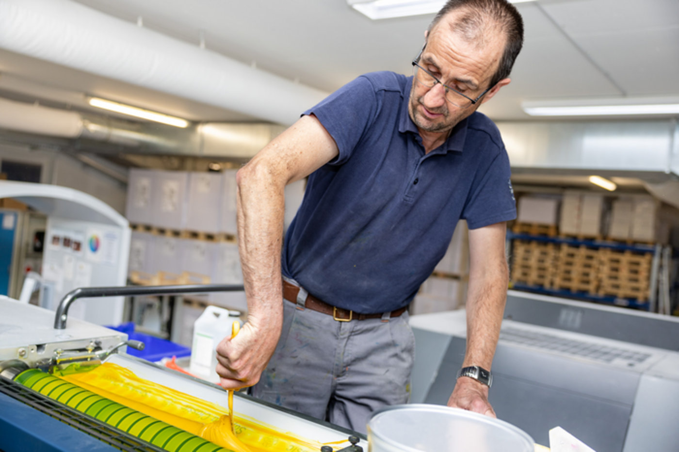Portrait Fotografie eines Drucktechnologen, welcher mit Spachtel gelbe Farbe auf der Walze einer Offsett- Druckmaschine verteilt.