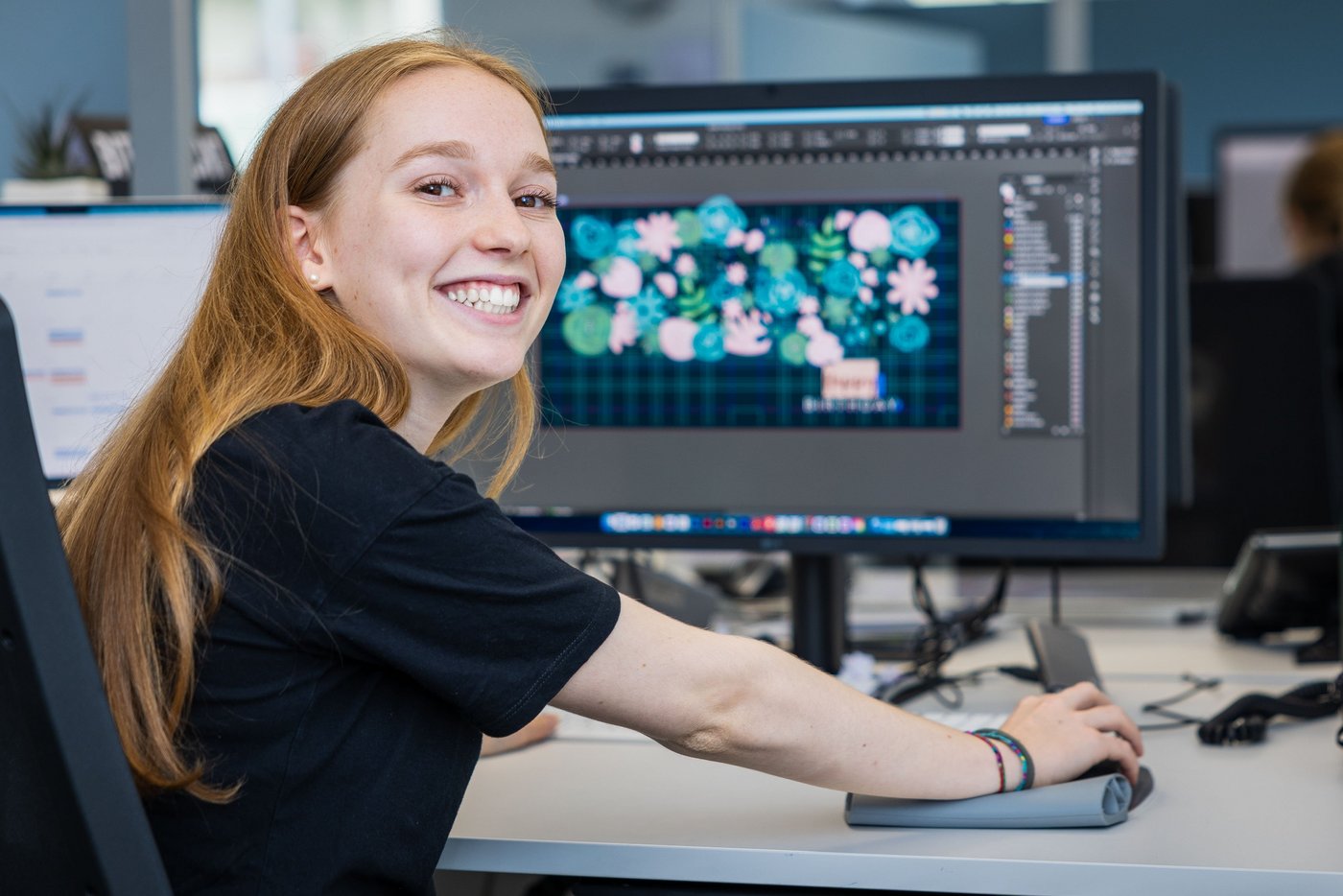 Portrait Fotografie einer Lernenden, welche am Bürotisch sitzt und eine Grafik erstellt und in die Kamera lacht.