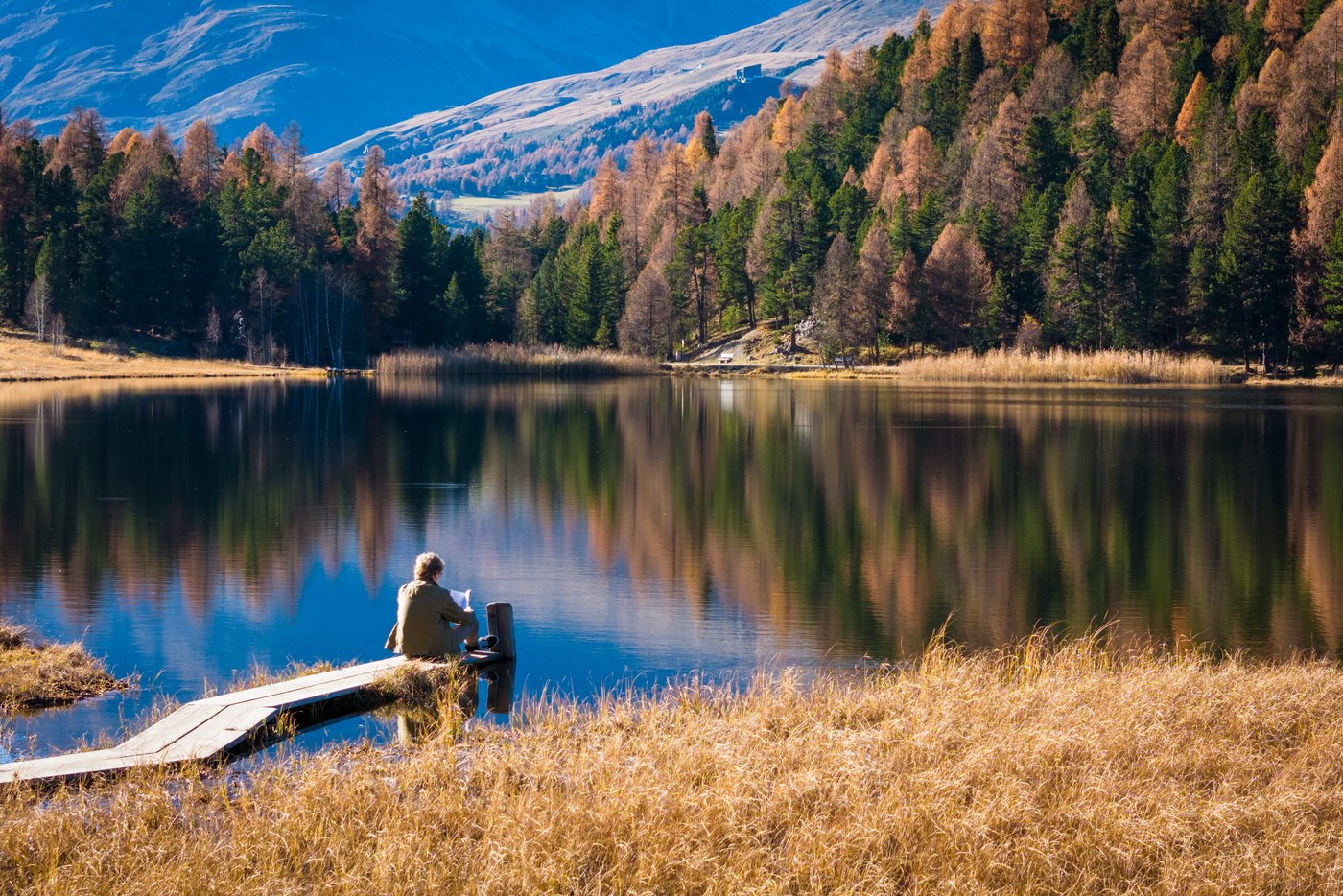 Landschaftsaufnahme des Stazersees im Engadin bei schönem Wetter. Eine Person sitzt auf dem Steg und liest ein Buch.